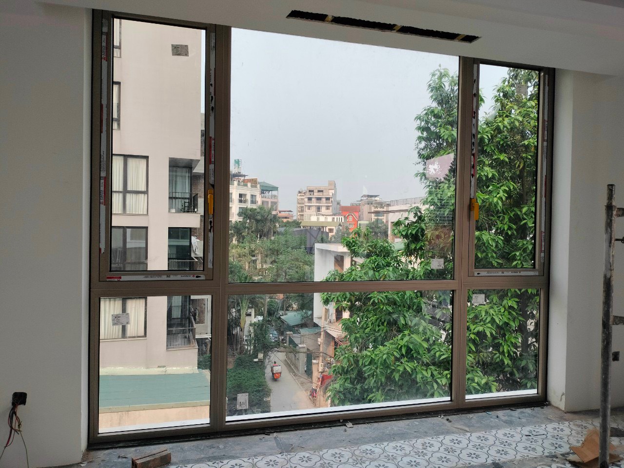 Cửa sổ nhôm kính khung nâu cỡ lớn nhìn ra phố và cây xanh trong căn hộ đang hoàn thiện